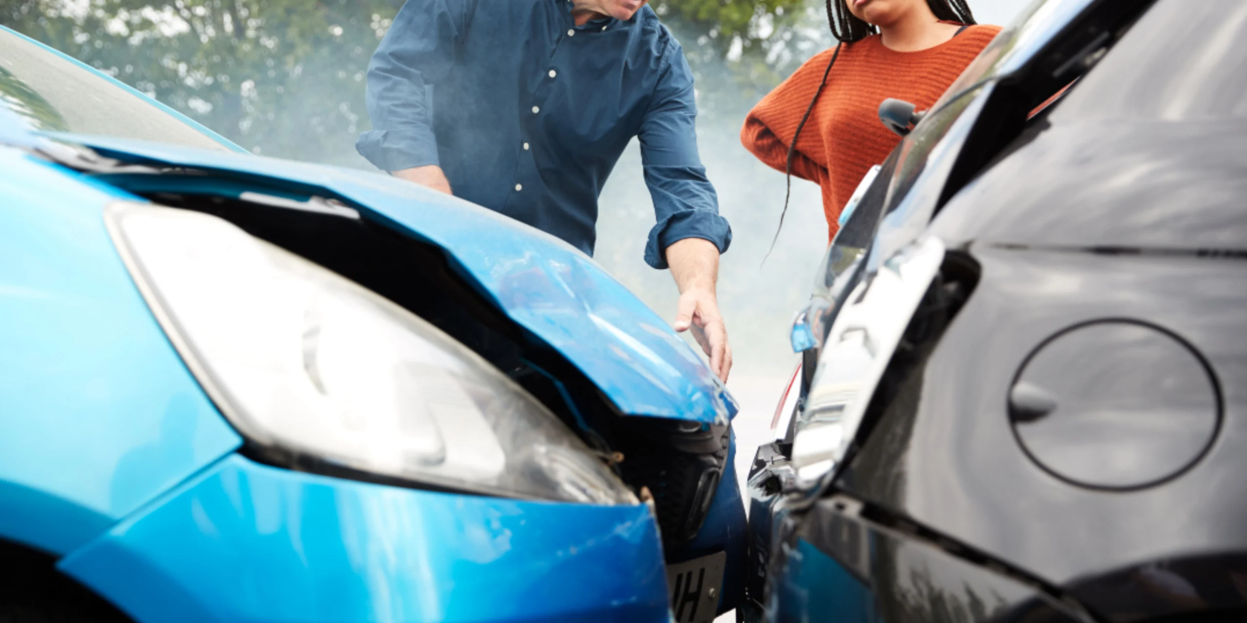 Two cars involved in a front end collision with visible damage to both vehicles while two people stand nearby inspecting the crash scene.