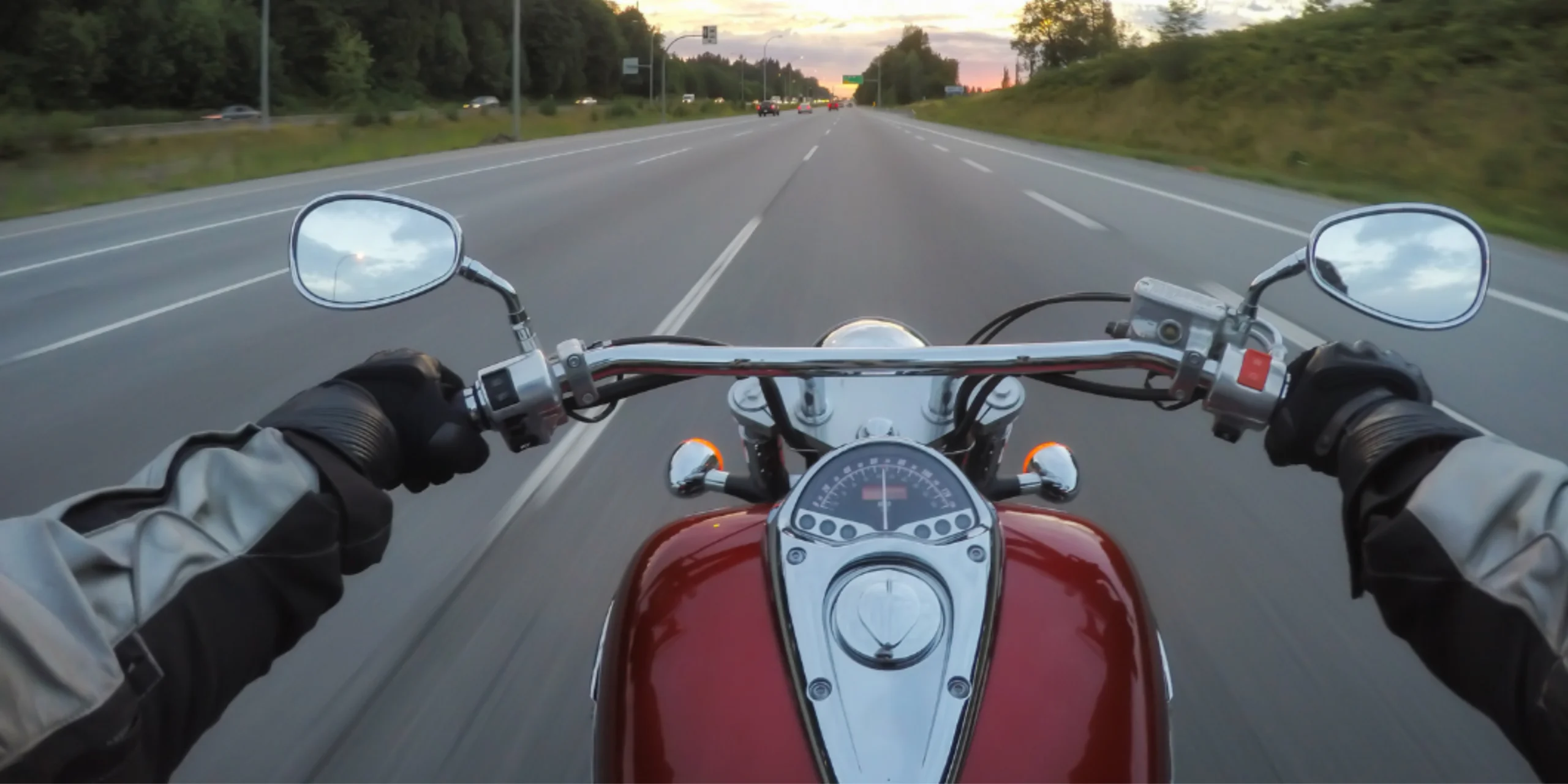 A first person view from a motorcyclist riding down a multi lane highway at sunset, with both hands on the handlebars and traffic ahead. The image highlights the rider's perspective and the potential risks motorcyclists face on busy roadways, illustrating the importance of safe riding practices and awareness to help prevent serious motorcycle accidents.