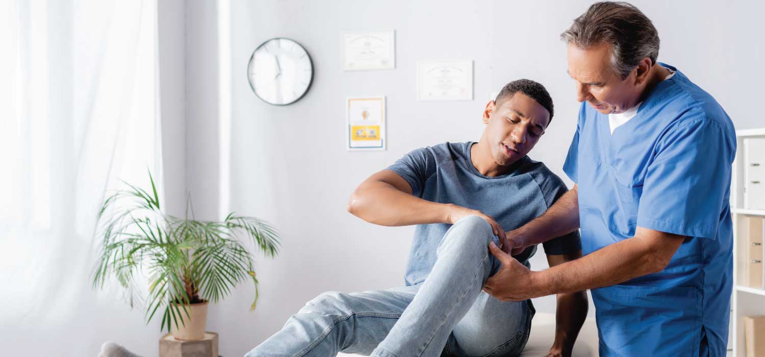 A male patient sits on an exam table and describes an injury in his left leg to a medical provider.