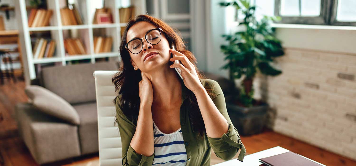 A woman holding her phone in one hand, holds her neck with the other and tries to stretch her neck out.